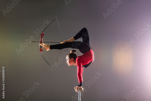 Young acrobat performing with archery bow on gymnastics bar