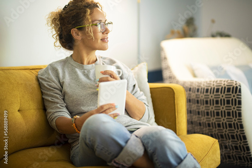 Beautiful young woman in casual clothes is using a digital tablet and smiling while sitting on couch at home. Yellow sofa. Smart modern female people enjoying technology and having relax leisure day photo