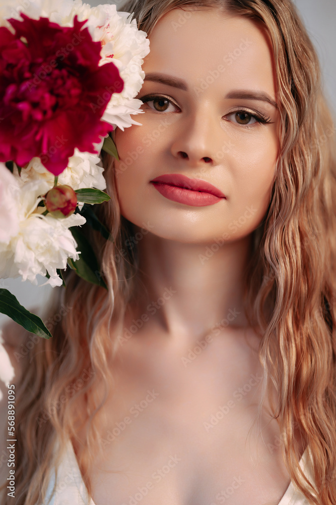 Attractive woman with long blond wavy hair holds her hands near her face. Around her are pink and white fresh flowers in a large beautiful bouquet of flowers. Artistic portrait with flowers.