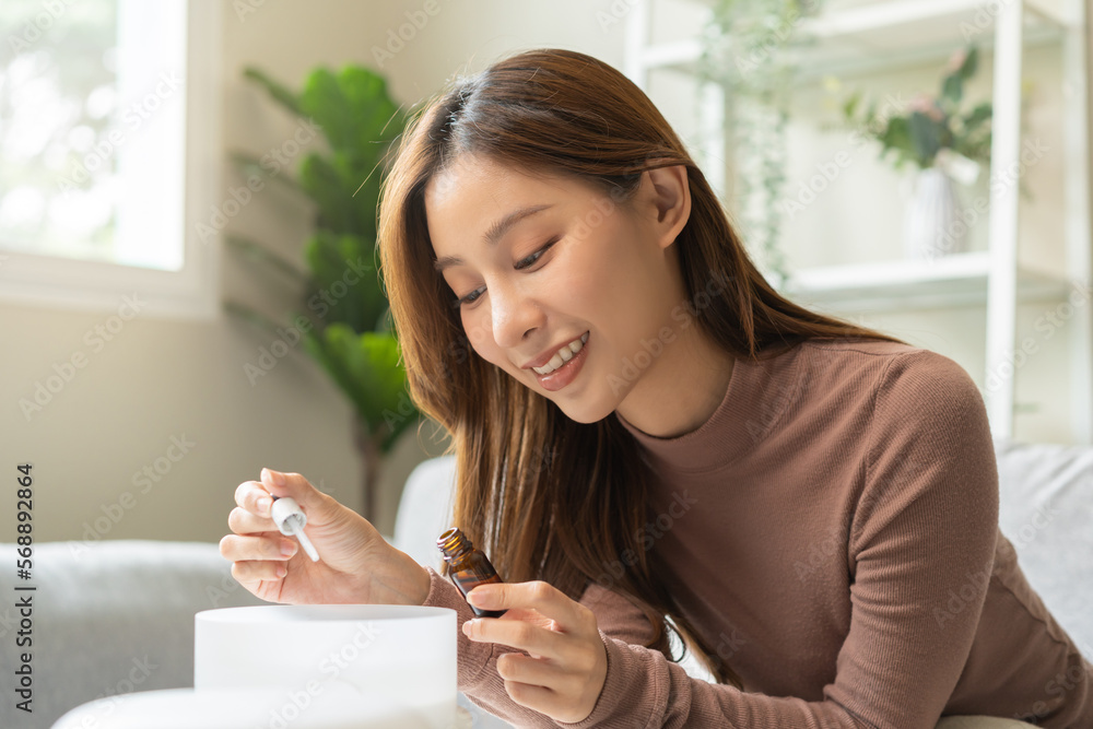 Happy asian young woman, girl hand holding bottle of essential and drop oil into air humidifier, enjoy aromatherapy steam scent diffuser comfortable while rest in living room, apartment at home. Happy asian young woman, girl hand holding bottle of essential and drop oil into air humidifier, enjoy aromatherapy steam scent diffuser comfortable while rest in living room, apartment at home.