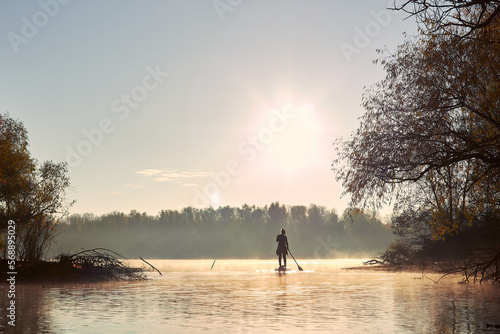 Rear view of woman paddling on stand up paddle board (SUP) at autumn misty river near shore with trees