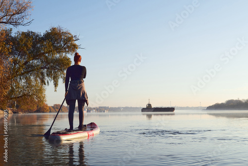 Rear view of woman paddle on stand up paddle board (SUP) at the morning on quiet autumn Danube river. Cargo ship on the horizon