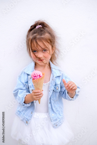 A girl in blue denim jacket and white skirt eats ice cream in waffle cone. Child showing success gesture, thumb up with hands, saying well done. The girl's curly hair is fluttering in the wind
