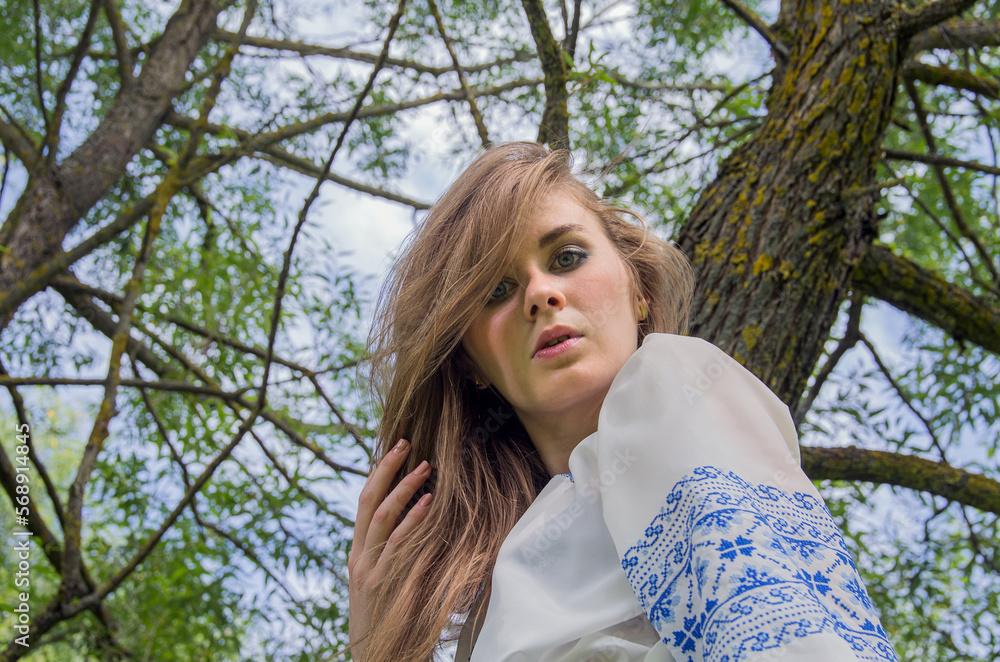 Naklejka premium A girl in a long blue skirt and an embroidered shirt among the green meadow and marsh grass and willow trees.
