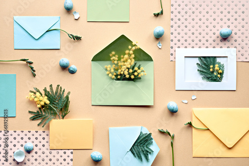 Easter background with quail eggs. Mimosa and freesia flowers  mint green envelopes and color paper on yellow background. Flat lay  overhead  knolling  neatly arranged springtime decor.