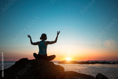 A woman does yoga, meditating by the ocean during sunset.