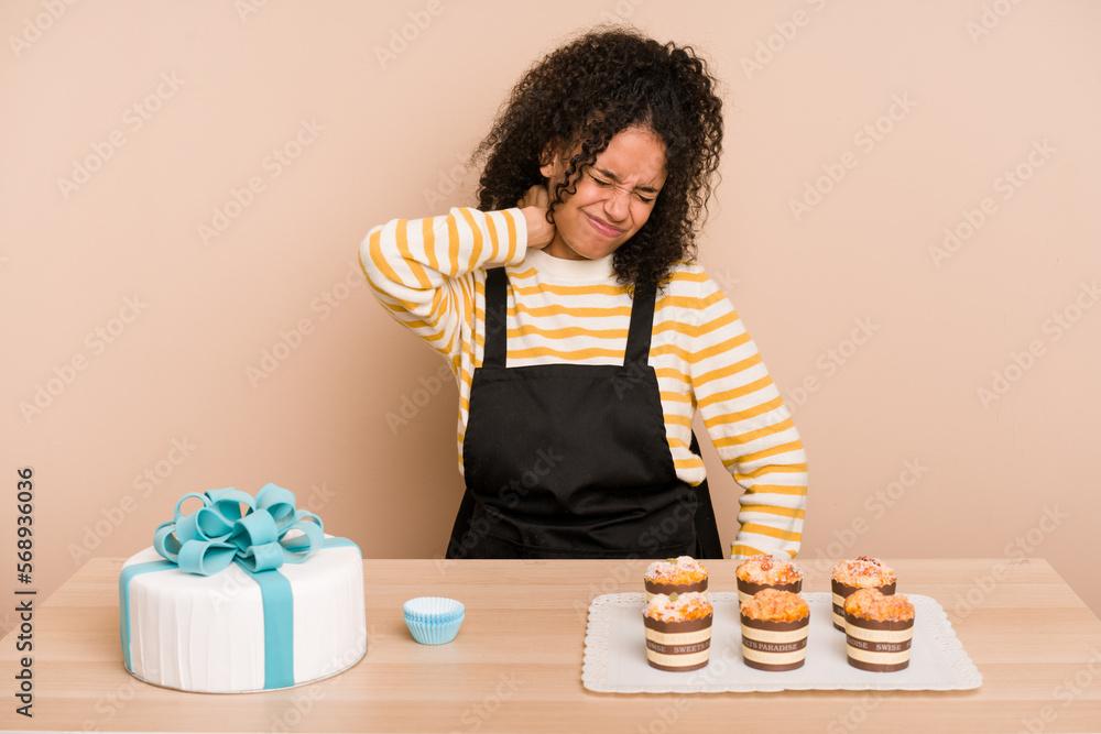 Young african american woman preparing a sweet cake and muffins on a table having a neck pain due to stress, massaging and touching it with hand. Young african american woman preparing a sweet cake and muffins on a table having a neck pain due to stress, massaging and touching it with hand.
