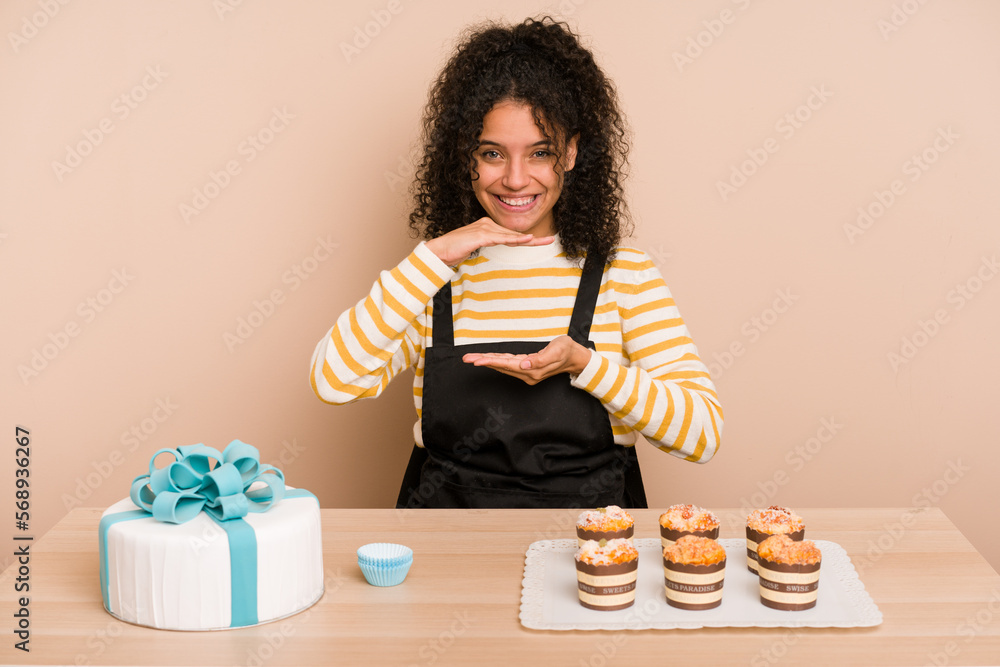 Young african american woman preparing a sweet cake and muffins on a table holding something with both hands, product presentation.