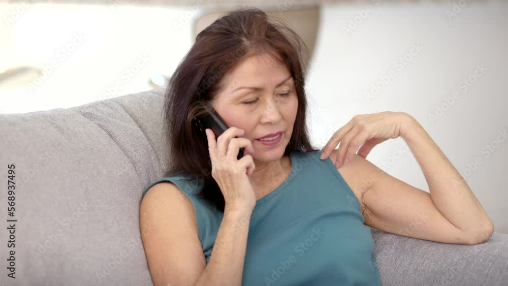 Beautiful smiling woman talking on cellphone sitting on comfortable couch in leisure home living room. Mature asian has healthy lifestyle. Beautiful smiling woman talking on cellphone sitting on comfortable couch in leisure home living room. Mature asian has healthy lifestyle.