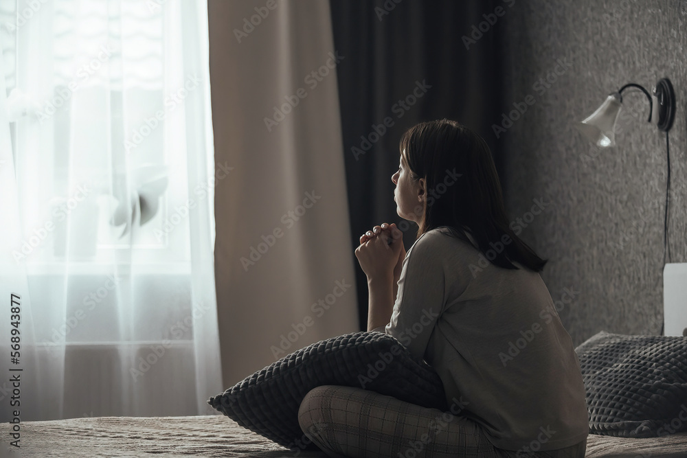 Naklejka premium A sad young woman in pajamas with seasonal affective disorder or depression sits alone on the bed and looks out the window. The concept of winter depression due to lack of sunlight