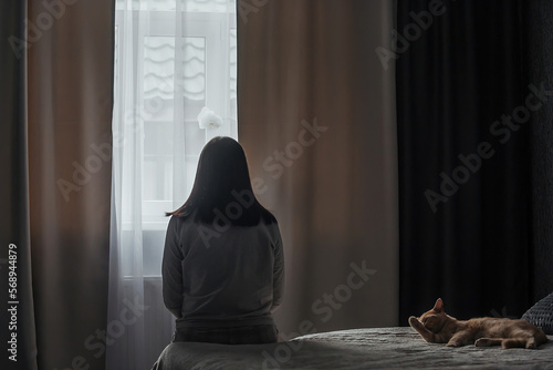 Фотография A sad young woman with seasonal affective disorder or depression sits alone on the bed and looks out the window, a domestic cat lies nearby