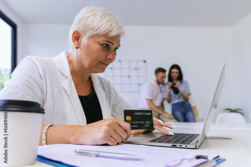 Senior woman consumer holding credit card and laptop buying online at home. Woman shipping online with credit card in hand. Women using laptop technology paying credit card shopping online