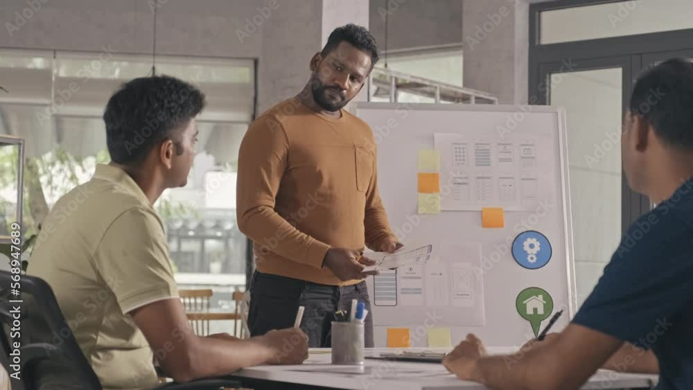 Indian UX designer standing by flipchart in office and giving presentation to team during business meeting Indian UX designer standing by flipchart in office and giving presentation to team during business meeting