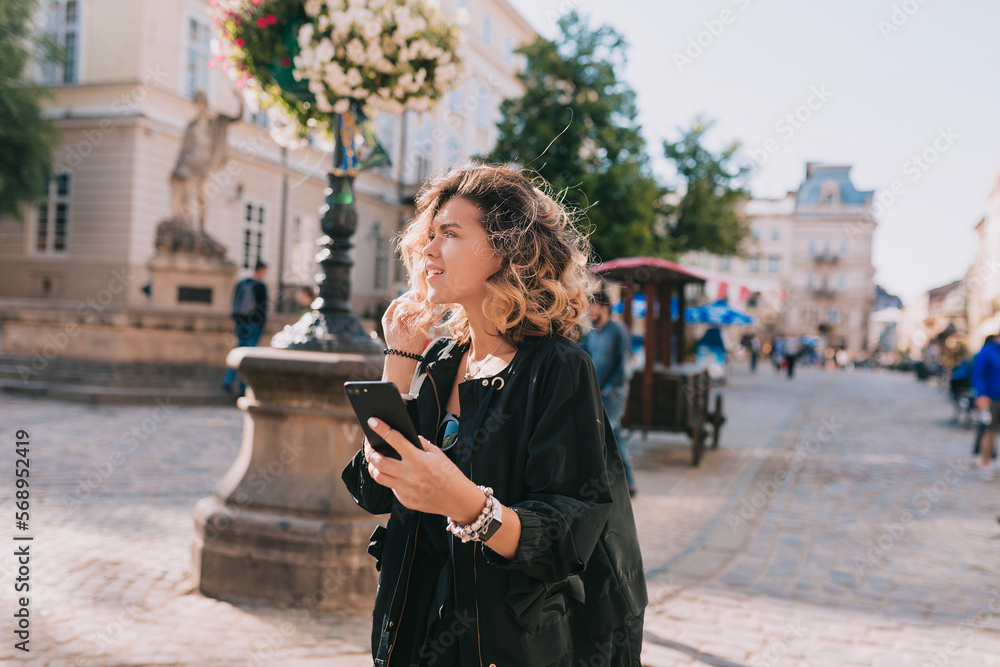 Romantic adorable happy girl with puffy hair wearing black jacket walking in city with smartphone in sunny warm day. Stylish girl exploring city in spring day. Romantic adorable happy girl with puffy hair wearing black jacket walking in city with smartphone in sunny warm day. Stylish girl exploring city in spring day.