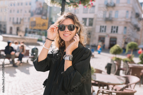 Cool stylish happy girl with curly light hair wearing black sunglasses posing to camera while talking on smartphone on city background in sunny warm day
