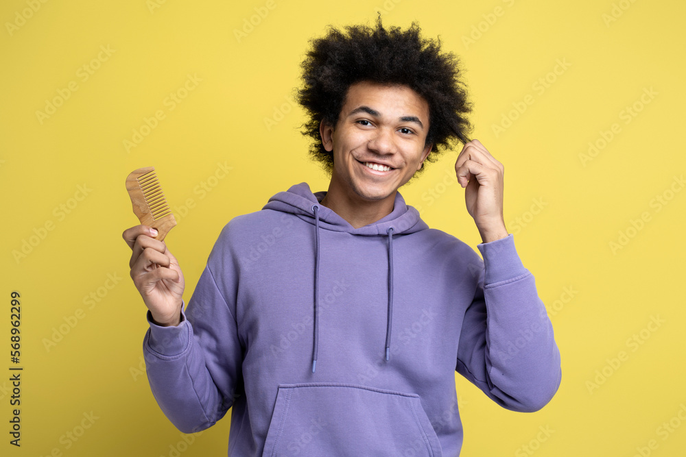 Smiling attractive African American man holding hair brush, touching his curly hair looking at camera isolated on yellow background. Hair care, morning routine concept Smiling attractive African American man holding hair brush, touching his curly hair looking at camera isolated on yellow background. Hair care, morning routine concept