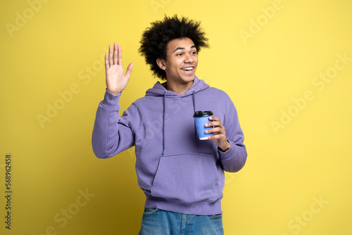 Smiling African American man wearing stylish hoodie, holding cup of coffee waving hand, isolated on yellow background 