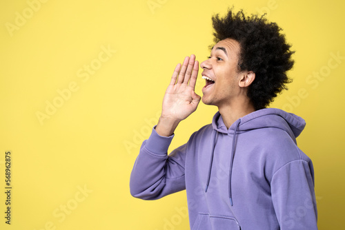 Young emotional African American man holding hand near face  screaming loud looking away isolated on yellow background. Happy curly haired guy doing sales announcement. Promotion  shopping concept 