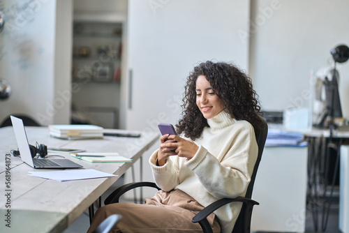 Young happy hispanic business woman office worker looking at smartphone using mobile cell phone technology, professional businesswoman executive working in office typing on cellphone sitting at desk.