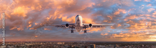 Airplane is flying in colorful sky over the city at sunset. Landscape with passenger airplane, skyline, blue sky with orange clouds. Aircraft is landing. Front view. Travel. Aerial view. Transport