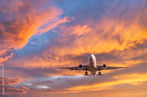 Airplane is flying in colorful sky at sunset. Landscape with passenger airplane, blue sky with orange and pink clouds. Aircraft is landing. Business and commercial. Travel. Aerial view. Transport