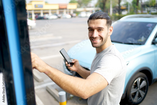 Cheerful happy Asian man using an EV charging application on smartphone to prepare vehicle charging and payment. Modern lifestyle of transportation with sustainability and sustainable energy.