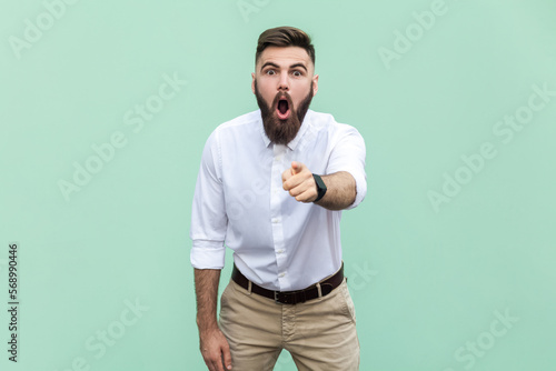 Surprised shocked bearded businessman wearing white shirt looking with big eyes and open mouth, pointing finger at camera, suspecting you. Indoor studio shot isolated on light green background.