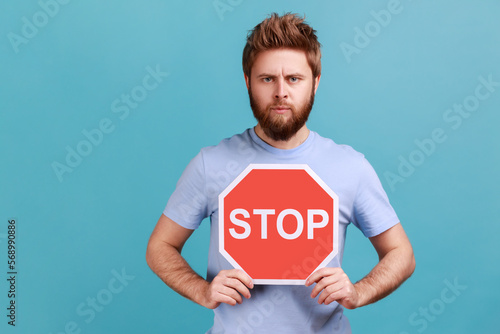 Portrait of handsome serious young adult bearded man holding octagonal Stop symbol, showing red traffic sign, warning of restricted area. Indoor studio shot isolated on blue background. © khosrork