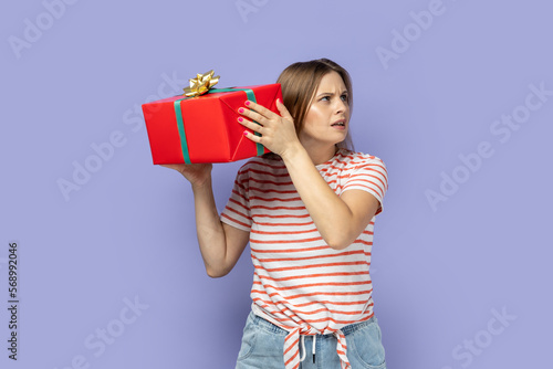 Curious beautiful blond woman wearing striped T-shirt holding and shaking red present box, being interested what gift her friends gave to her. Indoor studio shot isolated on purple background.