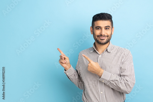 Satisfied bearded businessman pointing aside, showing blank copy space for idea presentation, commercial text, smiling happily, wearing striped shirt. Indoor studio shot isolated on blue background.
