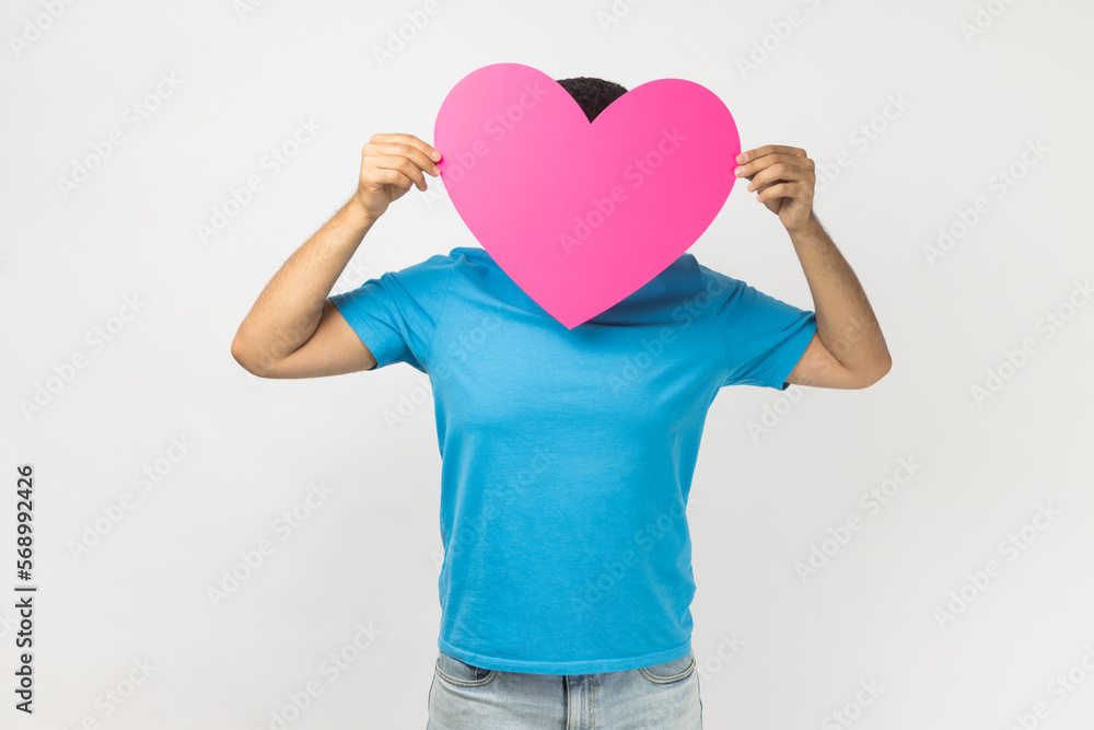 Portrait of romantic unknown anonymous man wearing blue T- shirt standing holding big pink heart in front his face, expressing fondness and love. Indoor studio shot isolated on gray background. Portrait of romantic unknown anonymous man wearing blue T- shirt standing holding big pink heart in front his face, expressing fondness and love. Indoor studio shot isolated on gray background.