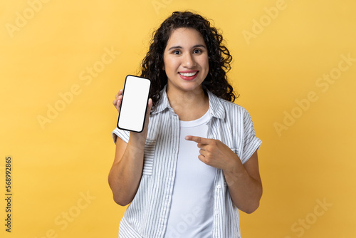 Portrait of smiling attractive young adult woman with dark wavy hair holding pointing at cell phone with empty display, copy space for advertisement. Indoor studio shot isolated on yellow background.