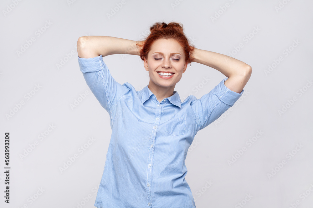 Portrait of beautiful attractive satisfied ginger woman wearing blue shirt standing with raised arms and closed eyes, smiling toothily. Indoor studio shot isolated on gray background. Portrait of beautiful attractive satisfied ginger woman wearing blue shirt standing with raised arms and closed eyes, smiling toothily. Indoor studio shot isolated on gray background.