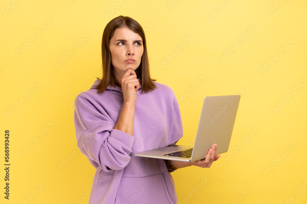 Pensive woman with working on laptop, holding chin, thinking about new project, looking away with thoughtful expression, wearing purple hoodie. Indoor studio shot isolated on yellow background. Pensive woman with working on laptop, holding chin, thinking about new project, looking away with thoughtful expression, wearing purple hoodie. Indoor studio shot isolated on yellow background.