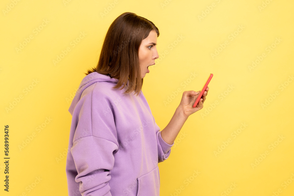 Side view of amazed excited woman reading message on smartphone, using mobile device for communication, surprised with app, wearing purple hoodie. Indoor studio shot isolated on yellow background.