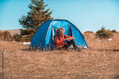 A man camping with a tent. He is surrounded by a beautiful mountain landscape  and the sun shines on his face  foreshadowing a beautiful and sunny day. The man looks satisfied and fulfilled