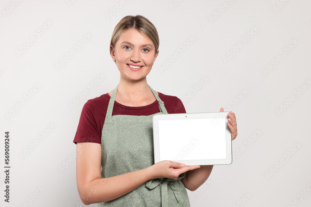 Beautiful young woman in clean apron with tablet on light grey background