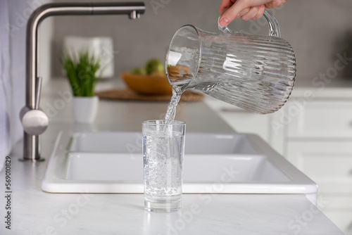 Woman pouring water from jug into glass at white countertop in kitchen, closeup