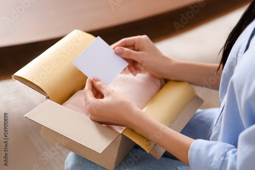 Woman holding blank greeting card near package with gift indoors, closeup