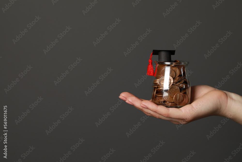 Woman holding glass jar of coins and graduation cap against dark grey background, closeup with space for text. Scholarship concept