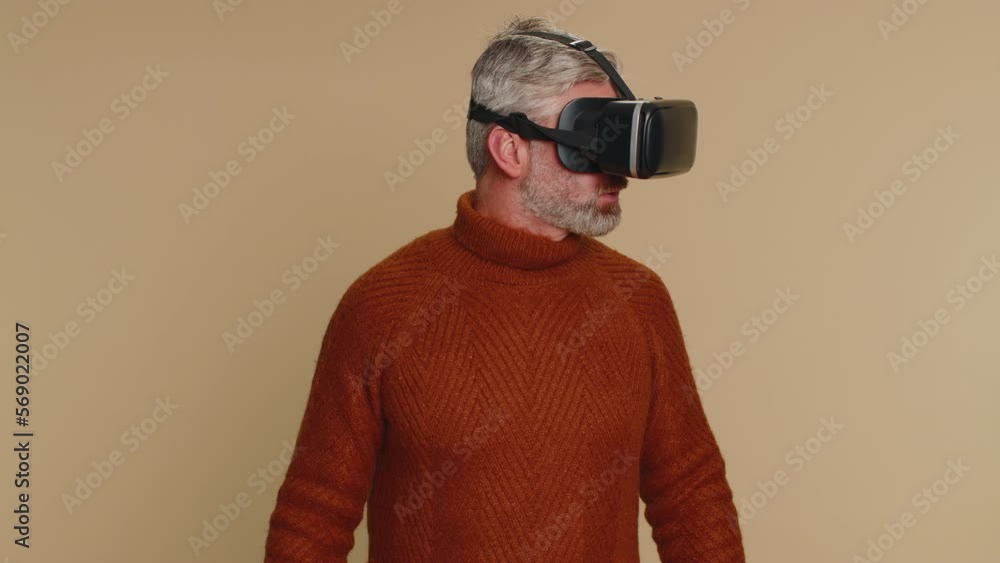 Middle-aged man using headset helmet to play simulation game app online. Watching virtual reality 3D 360 video content. One guy in VR goggles isolated on studio beige background. Futuristic technology Middle-aged man using headset helmet to play simulation game app online. Watching virtual reality 3D 360 video content. One guy in VR goggles isolated on studio beige background. Futuristic technology