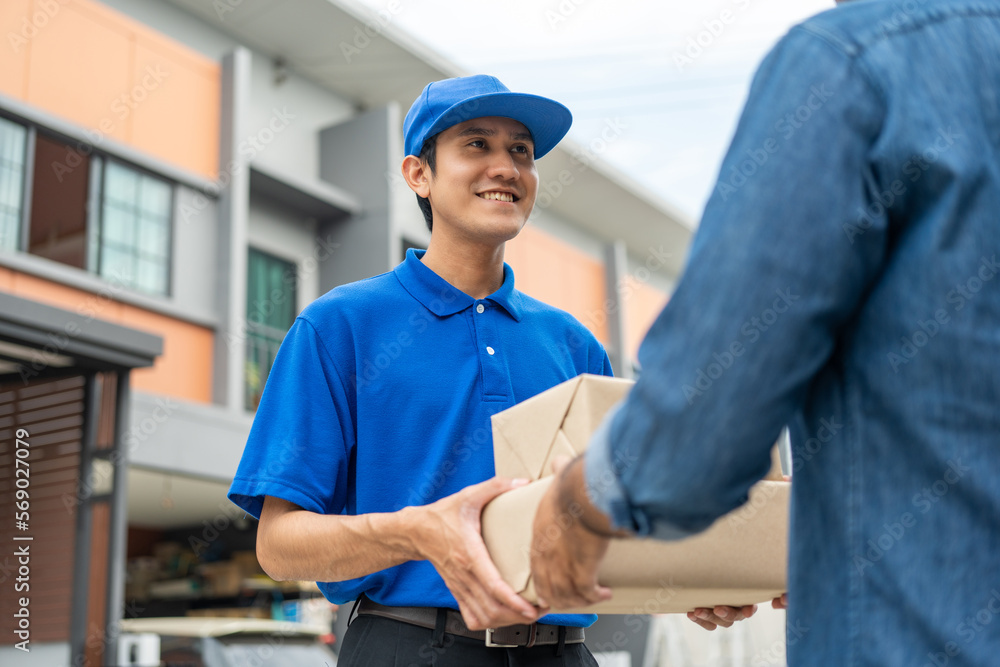 Fototapeta premium Asian delivery man with parcel in hand of blue uniform sending parcel to customer front of the house from shopping online with good service. Courier man send a package to destination.