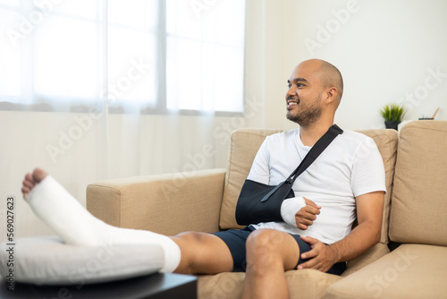 Happy man recovery from accident fracture broken bone injury with leg splints in cast neck splints collar arm splints sling support arm in living room. Social security and health insurance concept. © Chanakon