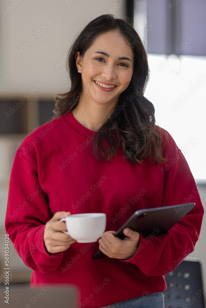 Image of young indian girl asian woman, company worker in dress red shirt, smiling and holding digital tablet, standing over office background Image of young indian girl asian woman, company worker in dress red shirt, smiling and holding digital tablet, standing over office background