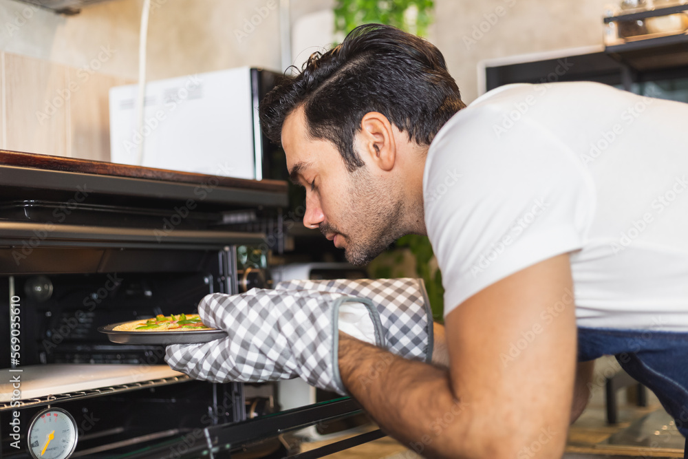 Smiling young Caucasian man in apron wearing oven mitt putting homemade pizza tray to the oven. Satisfied chef baking food at home. Italian delicious appetizer Smiling young Caucasian man in apron wearing oven mitt putting homemade pizza tray to the oven. Satisfied chef baking food at home. Italian delicious appetizer