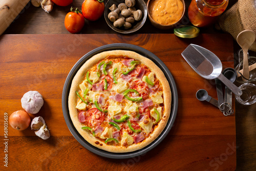 Top view of fresh baked homemade pizza on wooden table with ingredients and spices. Delicious Italian pizza is ready for deliver. Hawaiian Pizza with bell pepper bacon and pineapple.