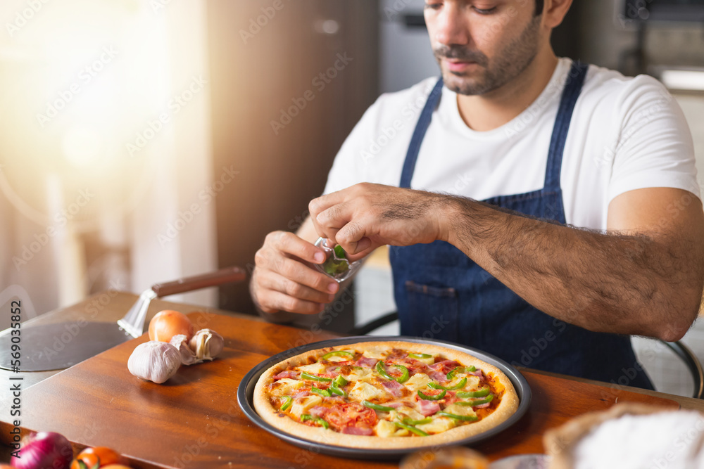 smiling young Caucasian man in apron and holding baking tray with fresh homemade pizza. Confident male chef holding cooked homemade pizza in kitchen. Italian delicious appetizer