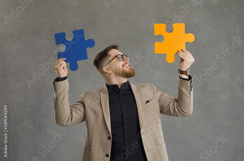 Happy creative guy in suit and modern hipster glasses joins together orange and blue jigsaw puzzle pieces standing isolated on grey background. Concept of finding good professional business solution photo