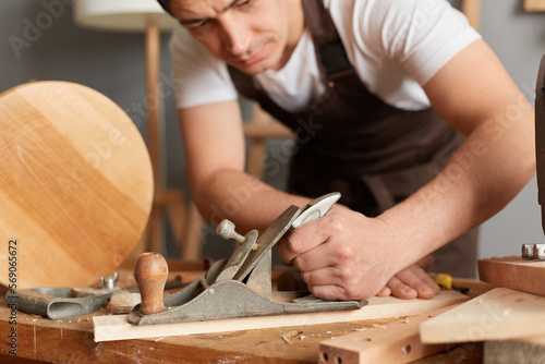 Closeup portrait of man carpenter wearing apron working with plane in wooden workshop, planing of a flat wooden surface, making furniture in his joinery.