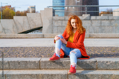 Young redhead woman in red coat yellow sweater and jeans sits on stairs in afternoon in sunny weather in the park in spring